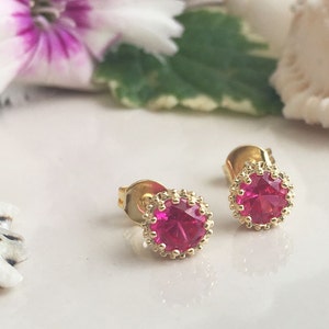 May include: A pair of gold stud earrings with a pink gemstone in the center of each earring. The gemstones are surrounded by a gold border.
