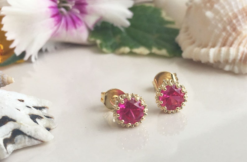May include: A pair of gold stud earrings with a pink gemstone in the center of each earring. The gemstones are surrounded by a gold border.