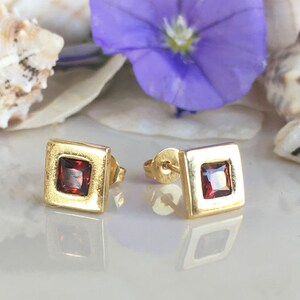 May include: A pair of gold square stud earrings with a red gemstone in the center of each earring.