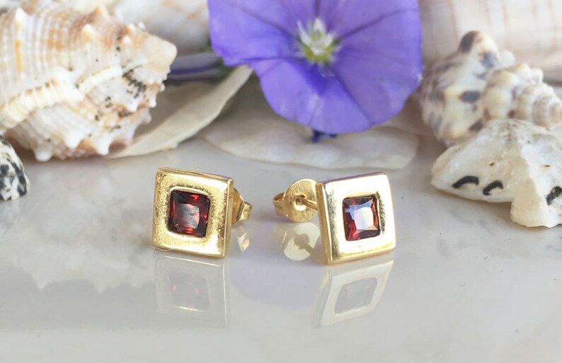 May include: A pair of gold square stud earrings with a red gemstone in the center of each earring.