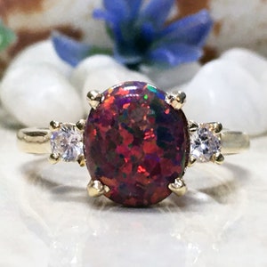 Può includere: Un anello d'oro con una grande pietra di opale rossa e due pietre trasparenti più piccole su entrambi i lati.