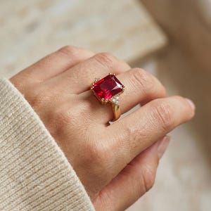 May include: A gold ring featuring a large, rectangular, vibrant red gemstone. The ring has a classic design with smaller, clear accent stones on either side of the main stone. The ring is shown on a hand.