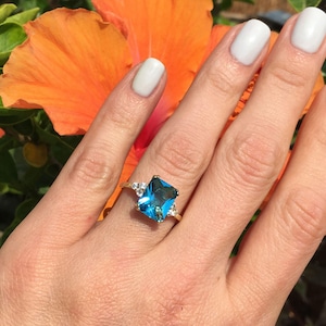 Könnte beinhalten: Ein goldener Ring mit einem großen, rechteckigen, leuchtend blauen Edelstein. Der Ring ist mit kleineren, klaren Edelsteinen auf beiden Seiten des Hauptsteins verziert. Der Ring ist vor dem Hintergrund einer orangefarbenen Blume platziert.