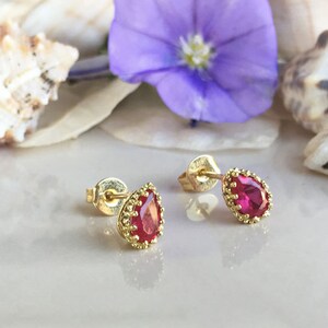 May include: A pair of gold earrings with red teardrop-shaped gemstones. The earrings have a delicate, ornate design.