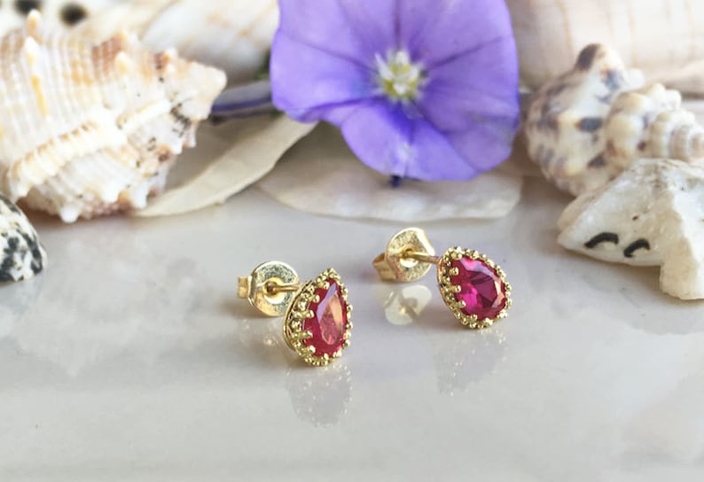 May include: A pair of gold earrings with red teardrop-shaped gemstones. The earrings have a delicate, ornate design.