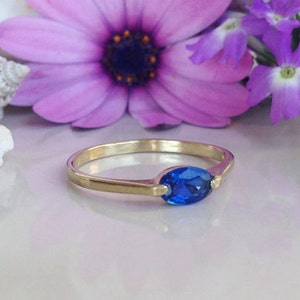 May include: A gold ring featuring a blue oval-shaped gemstone. The ring is displayed against a backdrop of purple flowers.