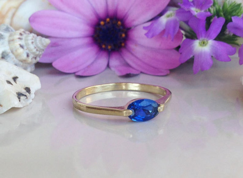 May include: A gold ring featuring a blue oval-shaped gemstone. The ring is displayed against a backdrop of purple flowers.