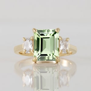 Puede incluir: Un anillo de oro con una gran piedra preciosa verde de talla esmeralda y dos piedras preciosas blancas rectangulares más pequeñas a cada lado.