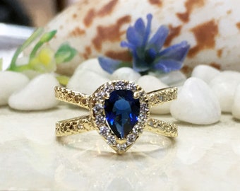 May include: A gold ring with a blue pear-shaped gemstone surrounded by smaller white gemstones. The ring has a criss-cross design.