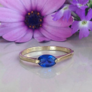 May include: A gold ring featuring a blue oval gemstone. The ring is displayed against a backdrop of seashells and purple flowers.