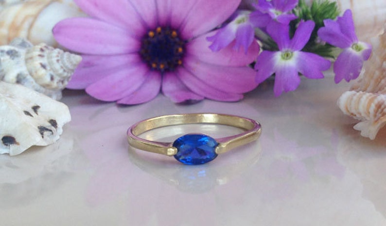 May include: A gold ring featuring a blue oval gemstone. The ring is displayed against a backdrop of seashells and purple flowers.