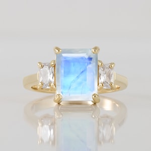 May include: A gold ring with a large, rectangular, iridescent blue moonstone and two smaller, rectangular, clear gemstones on either side.