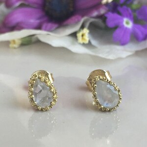 May include: A pair of gold earrings with a teardrop-shaped moonstone gemstone in each earring. The earrings have a delicate beaded border around the gemstone.