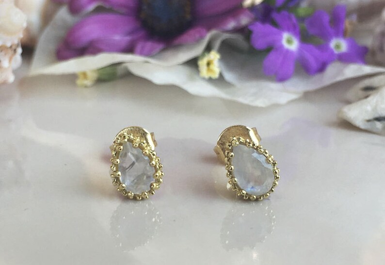 May include: A pair of gold earrings with a teardrop-shaped moonstone gemstone in each earring. The earrings have a delicate beaded border around the gemstone.