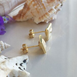 May include: A pair of gold square earrings with a white stone in the center. The earrings are on a white surface with seashells and flowers.