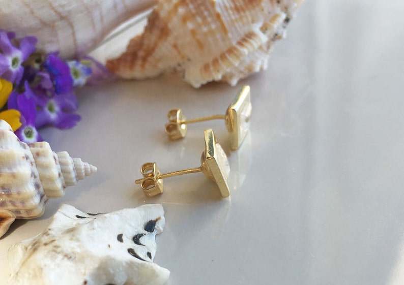 May include: A pair of gold square earrings with a white stone in the center. The earrings are on a white surface with seashells and flowers.