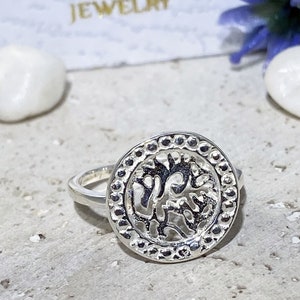 May include: A silver ring with a round, openwork design featuring a stylized tree motif. The ring has a beaded edge.
