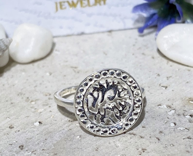 May include: A silver ring with a round, openwork design featuring a stylized tree motif. The ring has a beaded edge.