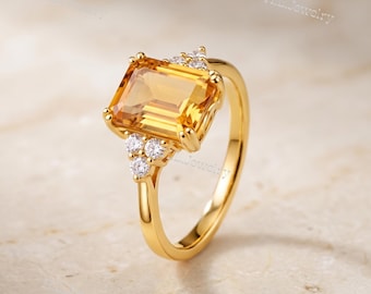 May include: A gold ring featuring a large, rectangular, emerald-cut yellow citrine gemstone as the center stone, flanked by small, round, clear accent stones on both sides. The band is polished gold, and the text H.L.Jewelry is visible.