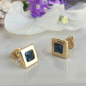 May include: A pair of gold square stud earrings with a blue gemstone in the center of each earring.