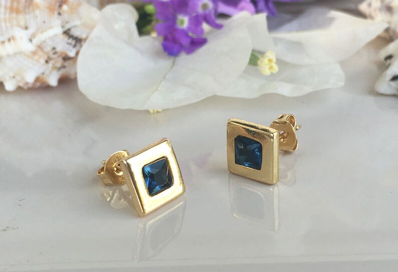 May include: A pair of gold square stud earrings with a blue gemstone in the center of each earring.