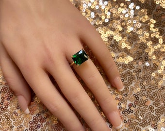 May include: A gold ring featuring a large, emerald-cut, deep green gemstone. The ring's band is a thin, polished gold. The gemstone is set with four prongs, showcasing its rectangular shape. The ring is displayed on a hand with manicured nails.