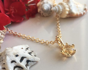 May include: A gold chain necklace with a small, round, light brown gemstone pendant. The pendant is set in a gold setting with a delicate, floral design. The necklace is photographed against a background of seashells.