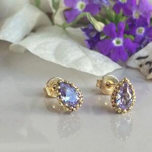 May include: A pair of gold earrings with a teardrop-shaped purple gemstone set in a gold bezel. The earrings have a delicate, ornate design.