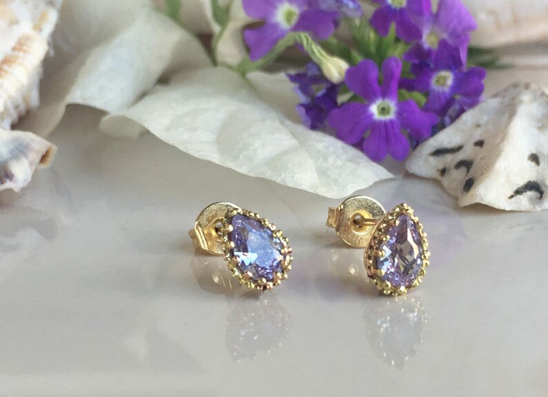 May include: A pair of gold earrings with a teardrop-shaped purple gemstone set in a gold bezel. The earrings have a delicate, ornate design.