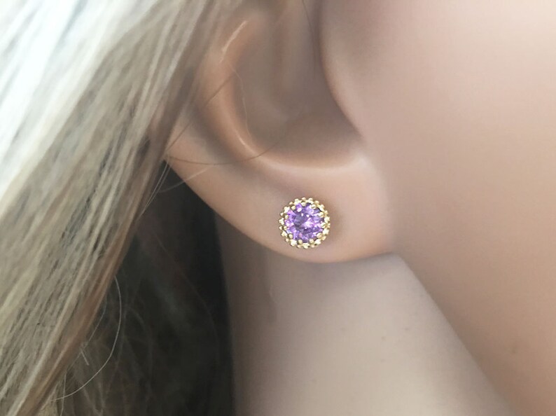May include: A pair of gold stud earrings with a small, round, purple gemstone in the center. The gemstone is surrounded by a halo of smaller, gold beads.