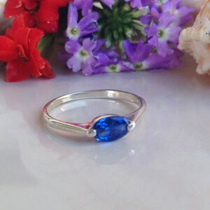 May include: A silver ring featuring a blue oval-shaped gemstone. The ring is displayed against a backdrop of seashells and flowers.