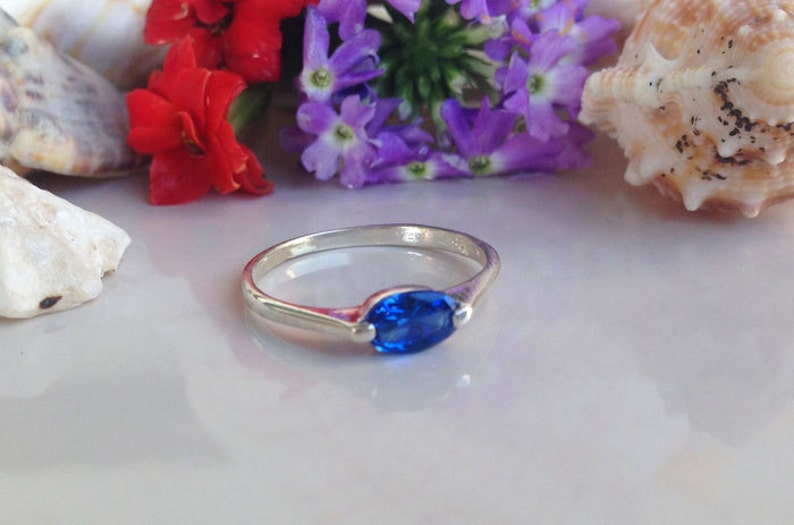 May include: A silver ring featuring a blue oval-shaped gemstone. The ring is displayed against a backdrop of seashells and flowers.