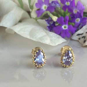 May include: A pair of gold earrings with a teardrop-shaped purple gemstone set in a gold bezel. The earrings have a delicate, ornate design.