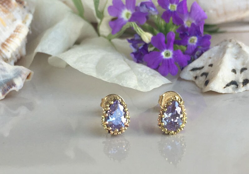 May include: A pair of gold earrings with a teardrop-shaped purple gemstone set in a gold bezel. The earrings have a delicate, ornate design.
