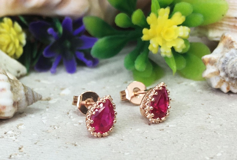 May include: A pair of rose gold earrings with teardrop-shaped red gemstones set in a delicate, ornate design.