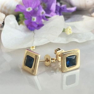 May include: A pair of gold square stud earrings with a dark blue square gemstone in the center of each earring.