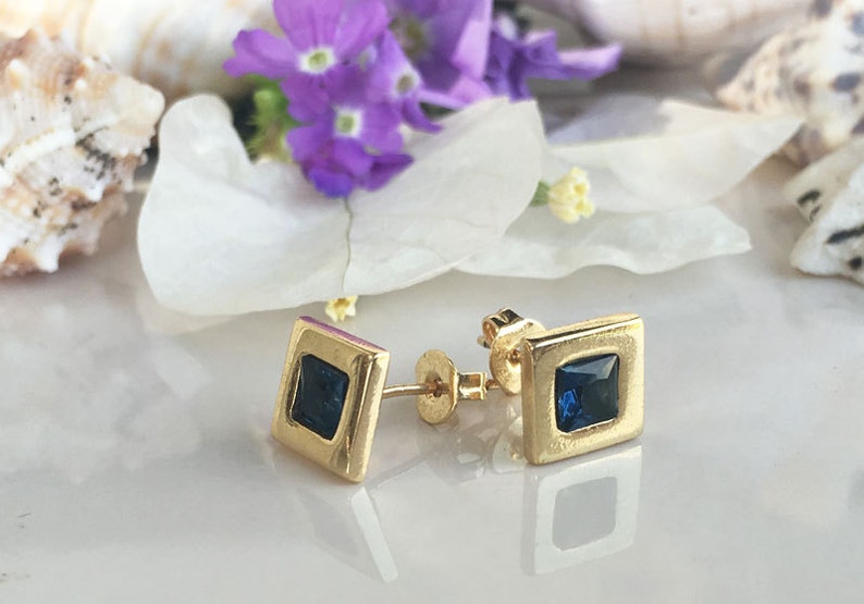 May include: A pair of gold square stud earrings with a dark blue square gemstone in the center of each earring.