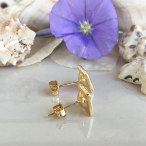 May include: A pair of gold-toned stud earrings with a geometric design. The earrings are shown on a white surface with seashells and a purple flower.