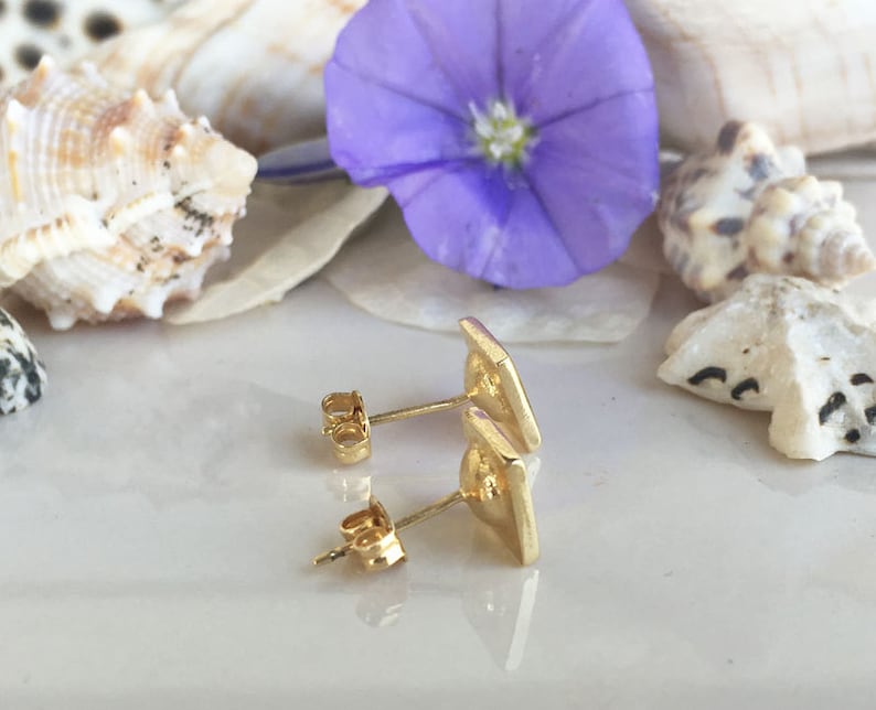 May include: A pair of gold-toned stud earrings with a geometric design. The earrings are shown on a white surface with seashells and a purple flower.