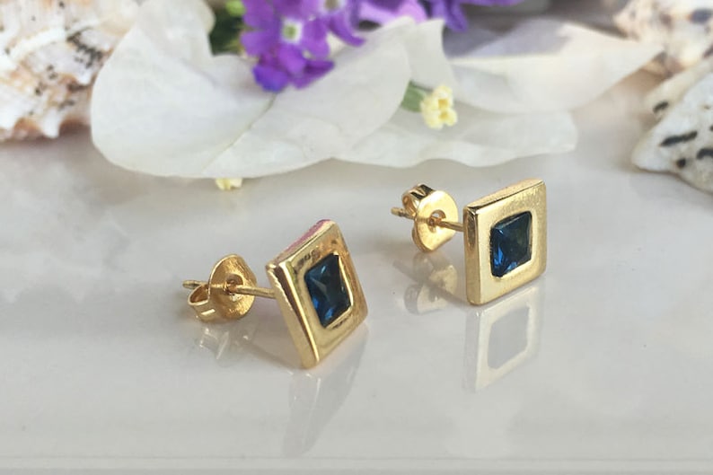 May include: A pair of gold square stud earrings with a blue gemstone in the center of each earring.