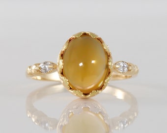 Puede incluir: Un anillo de oro con una gran piedra ovalada de ámbar engastada en un engaste de oro decorativo. Dos piedras pequeñas transparentes están engastadas a cada lado de la piedra principal.