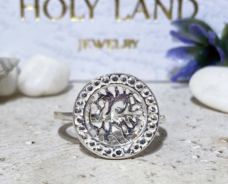 May include: A silver ring with a circular design featuring a tree of life motif. The ring has a beaded edge.