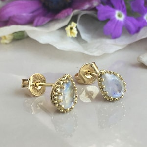 May include: A pair of gold earrings with teardrop-shaped moonstone gemstones. The earrings have a delicate gold beaded border around the stones.