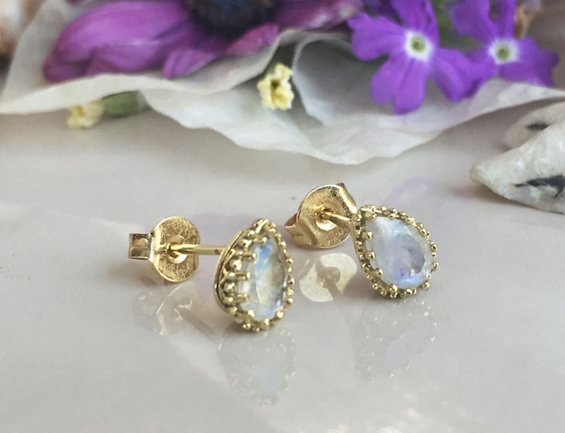 May include: A pair of gold earrings with teardrop-shaped moonstone gemstones. The earrings have a delicate gold beaded border around the stones.