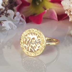 May include: A gold ring with a circular design featuring a cutout pattern. The ring is on a white surface.