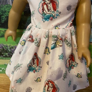 May include: A white sleeveless dress with a pink and blue pattern of Ariel the Little Mermaid and sea creatures. The dress has a gathered skirt and is perfect for a little girl.