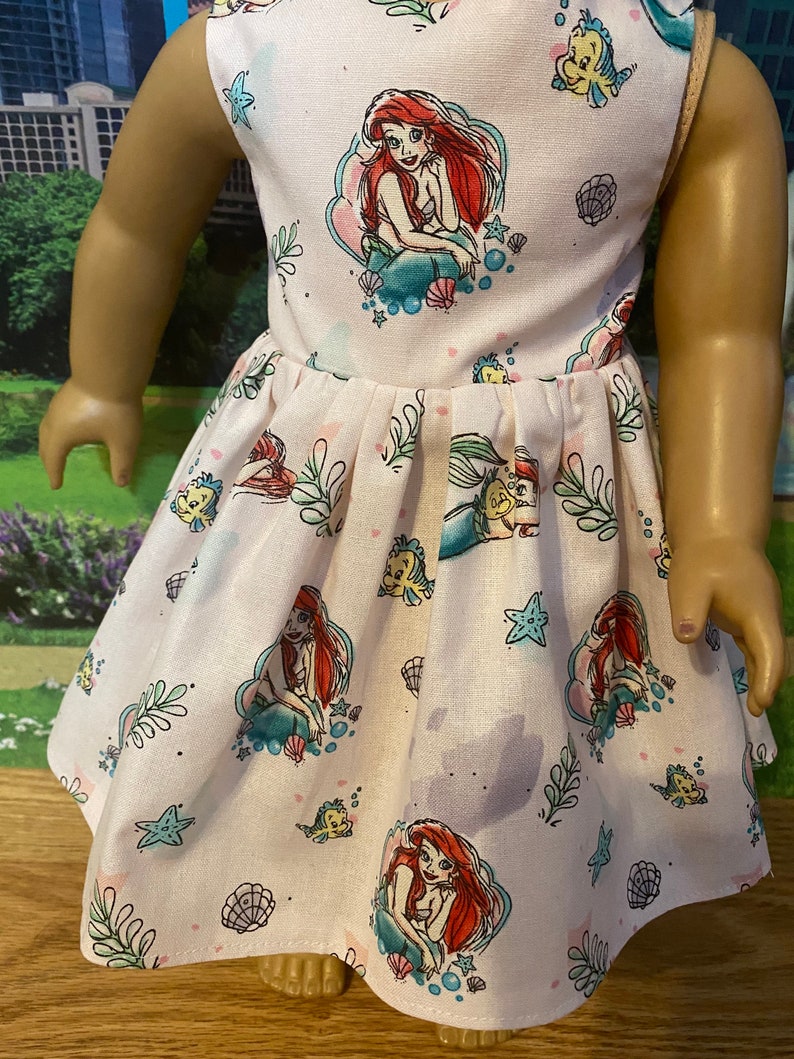 May include: A white sleeveless dress with a pink and blue pattern of Ariel the Little Mermaid and sea creatures. The dress has a gathered skirt and is perfect for a little girl.