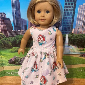 May include: A pink sleeveless dress with a pattern of Ariel from The Little Mermaid, seashells, and seahorses. The dress is made for a doll.