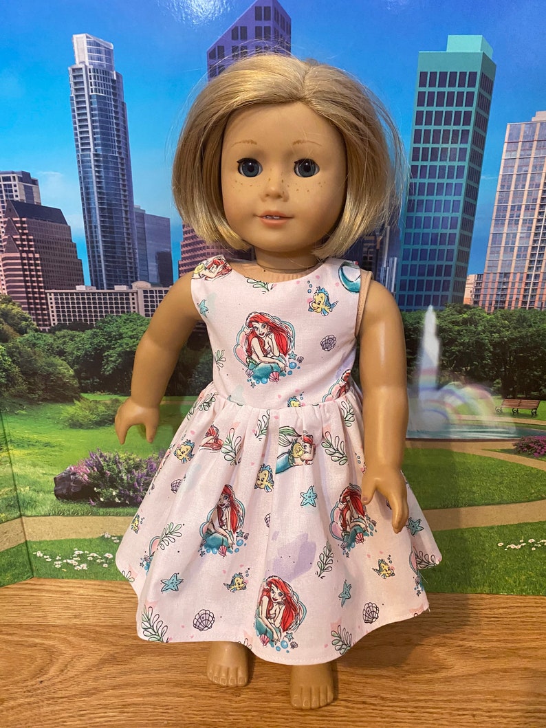 May include: A pink sleeveless dress with a pattern of Ariel from The Little Mermaid, seashells, and seahorses. The dress is made for a doll.
