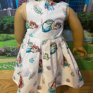May include: A pink sleeveless dress with a mermaid print. The dress features Ariel, Flounder, and Sebastian from the Disney film The Little Mermaid.